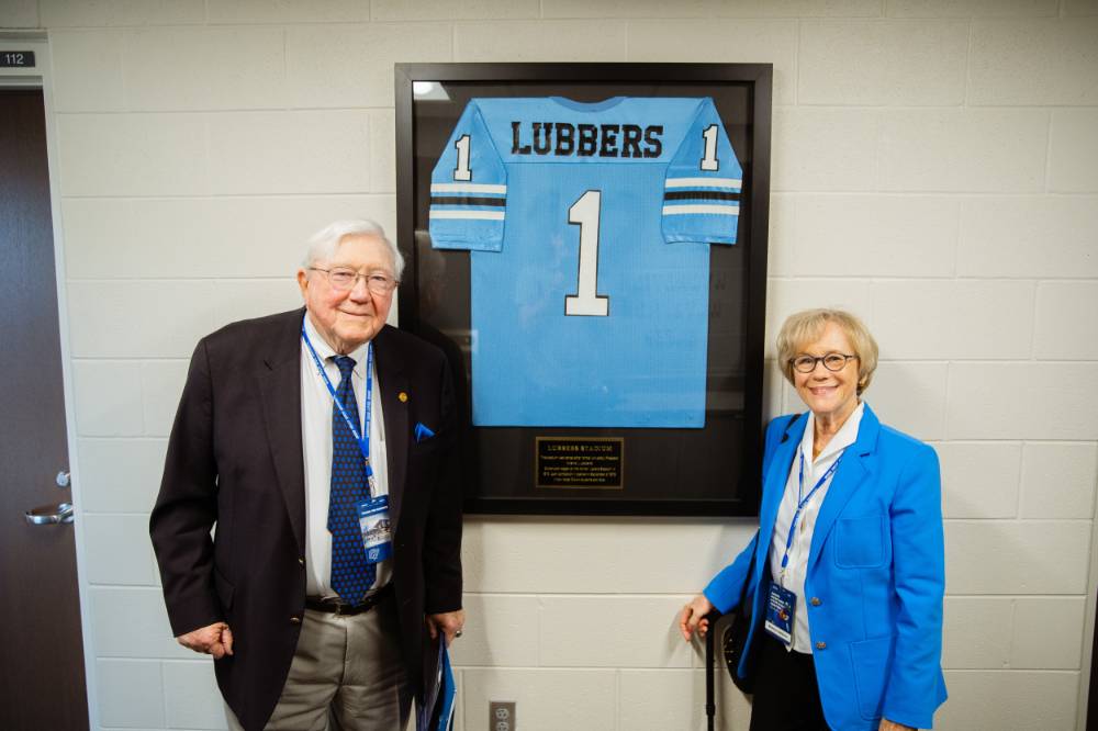 Don and Nancy Lubbers posing in front of a framed Lubbers jersey in the Jamie Hosford Football Center.